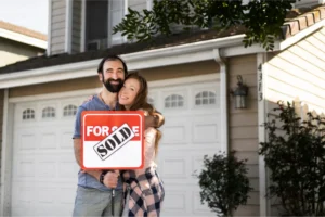 Happy couple standing in front of their new home, holding a 'For Sale' sign with a 'Sold' sticker.