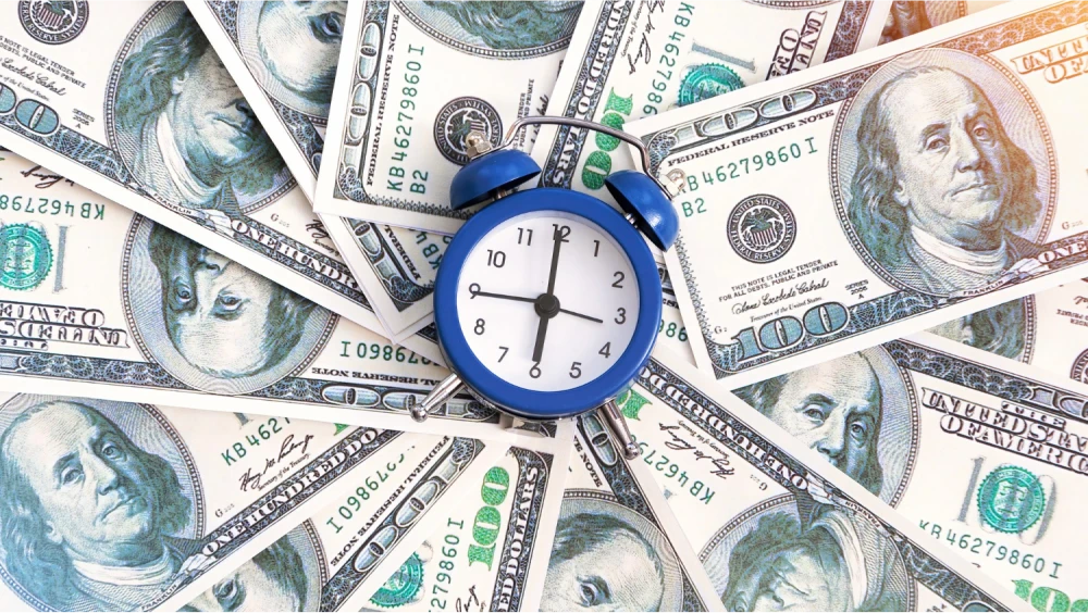 Alarm clock surrounded by a circle of US dollar bills, representing the concept of time and money.