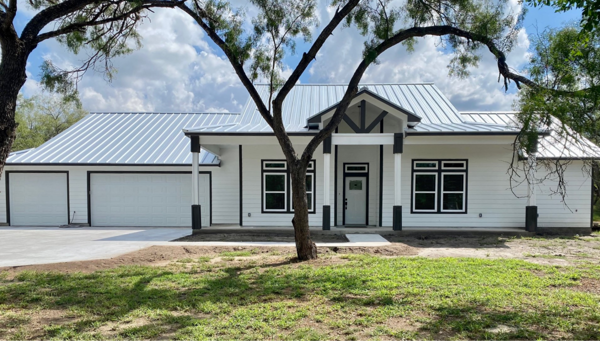 A white farmhouse with a metal roof, three-car garage, and large windows surrounded by trees.